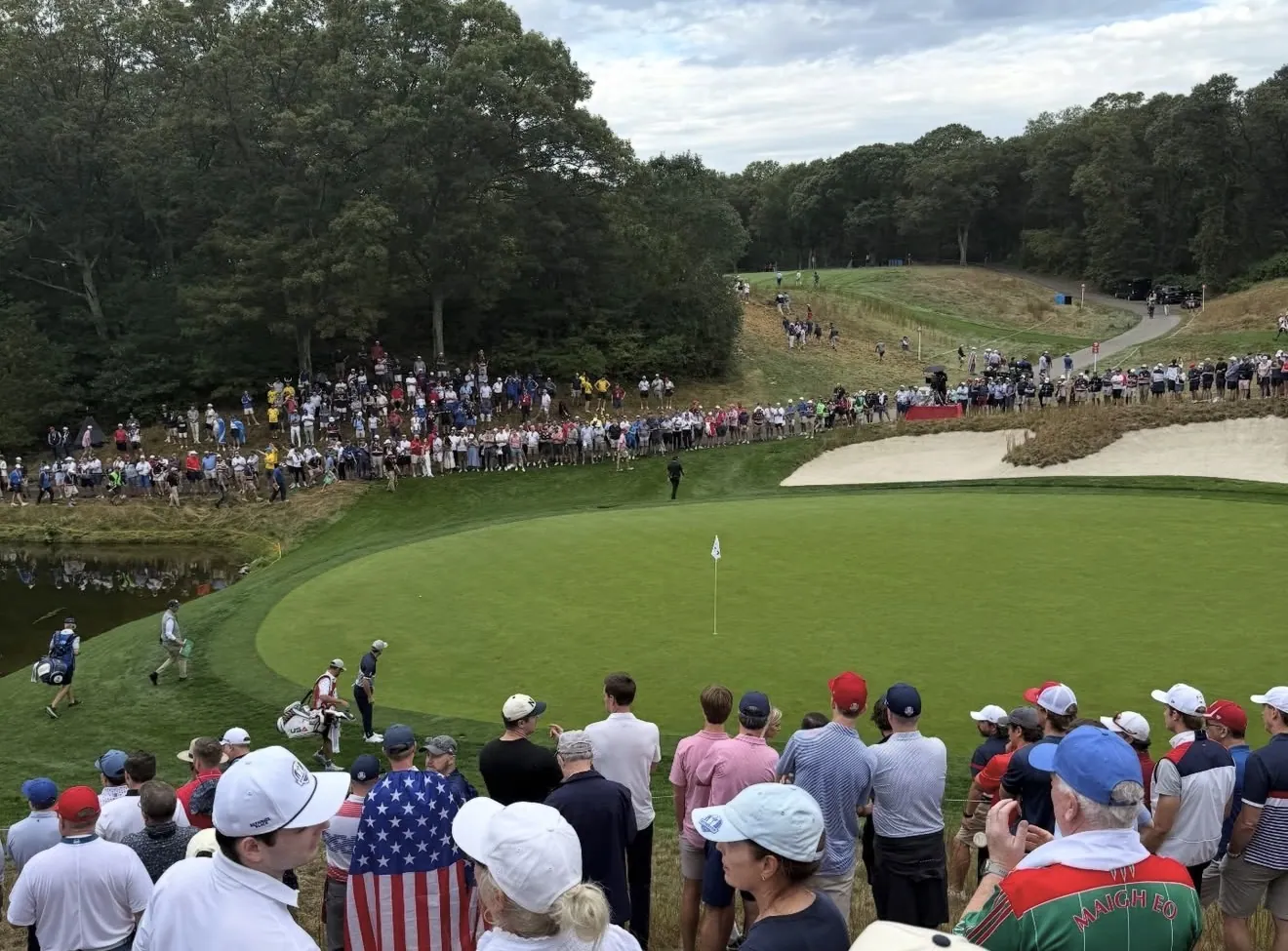 Ryder Cup 2025 crowd at Bethpage Black with a US flag held up and a USA caddie bag beside the green