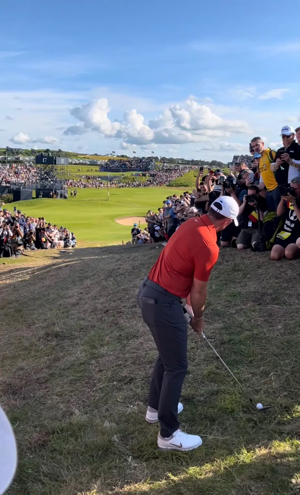 Golfer in red shirt playing a chip beside a large crowd of photographers at Royal Portrush